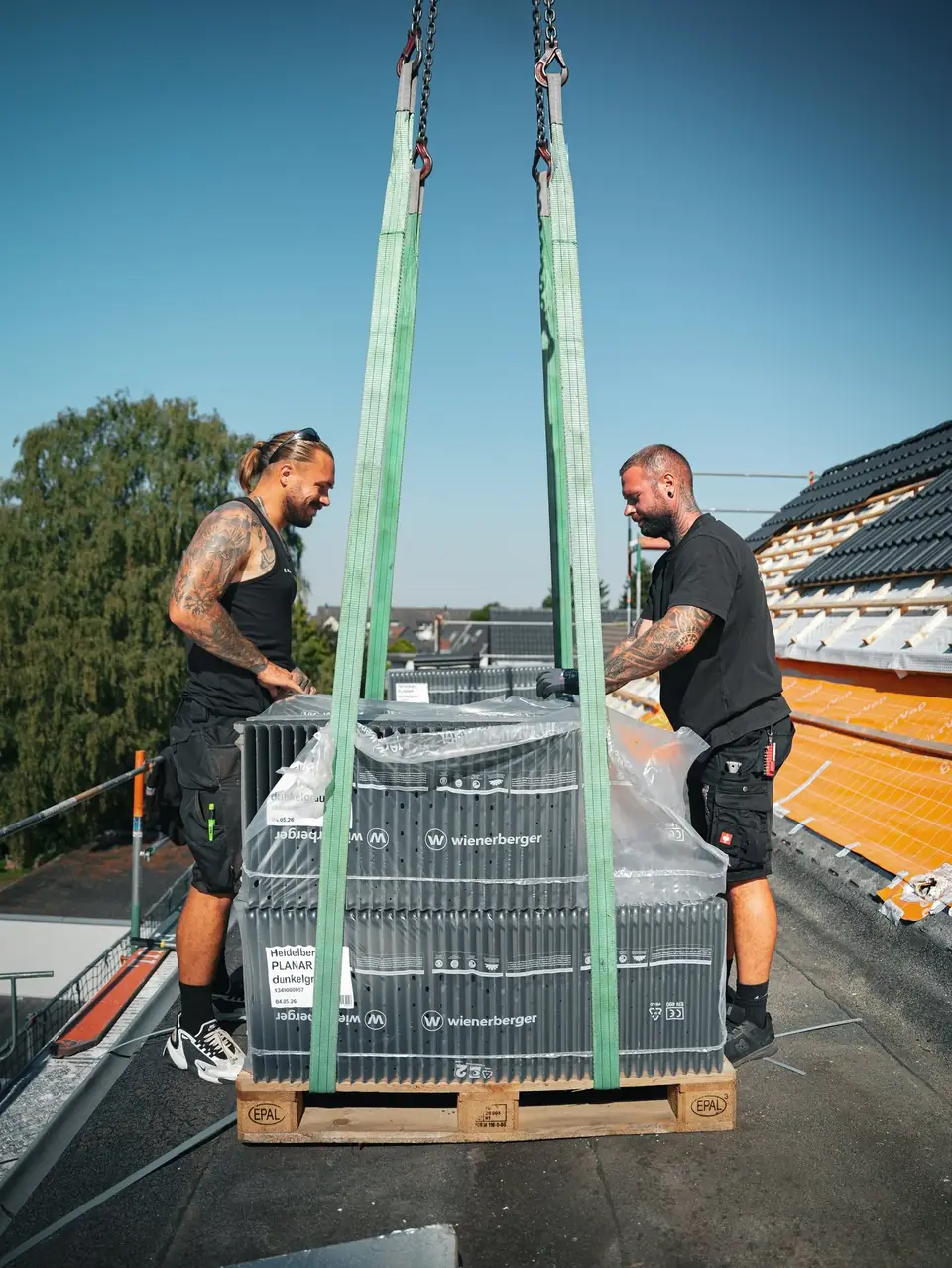 Zwei Dachdecker nehmen eine mit dem Kran angelieferte Palette Dachziegel auf dem Dach an